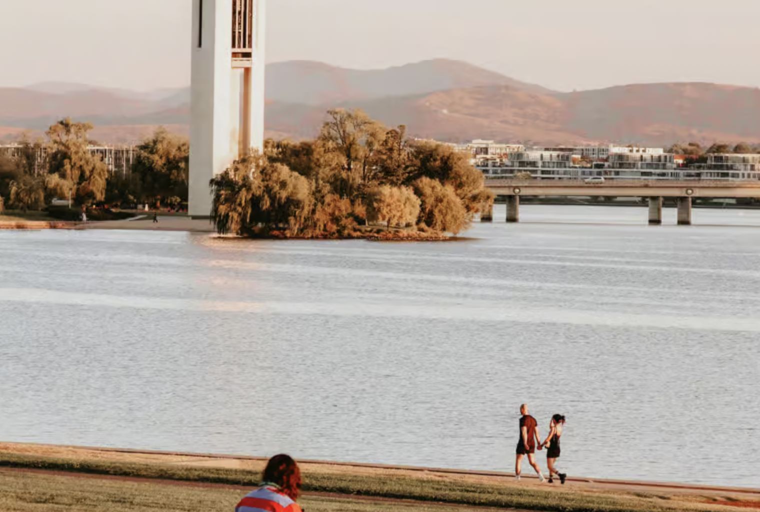 Couple walking along a lakeside at sunset with an islanded cluster of trees and a tall white tower, plus a distant bridge.